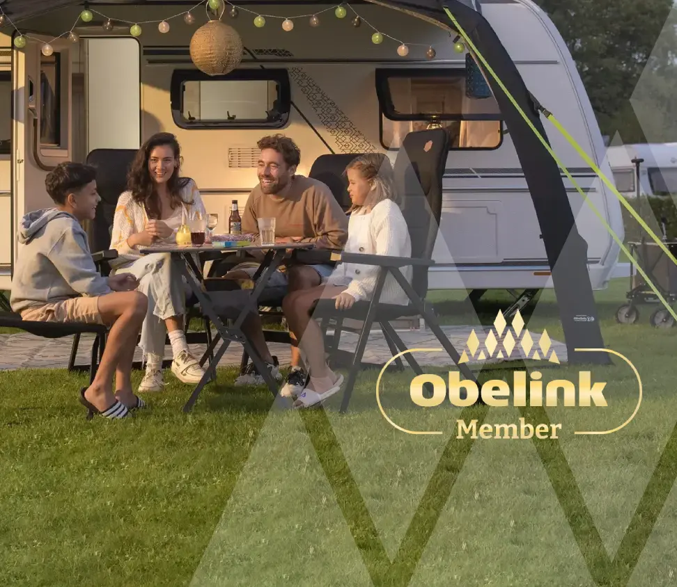 Sparen voor korting Caravan met luifel versierd met sfeerverlichting, mensen genieten samen aan een kampeertafel in de avondschemering op een grasveld