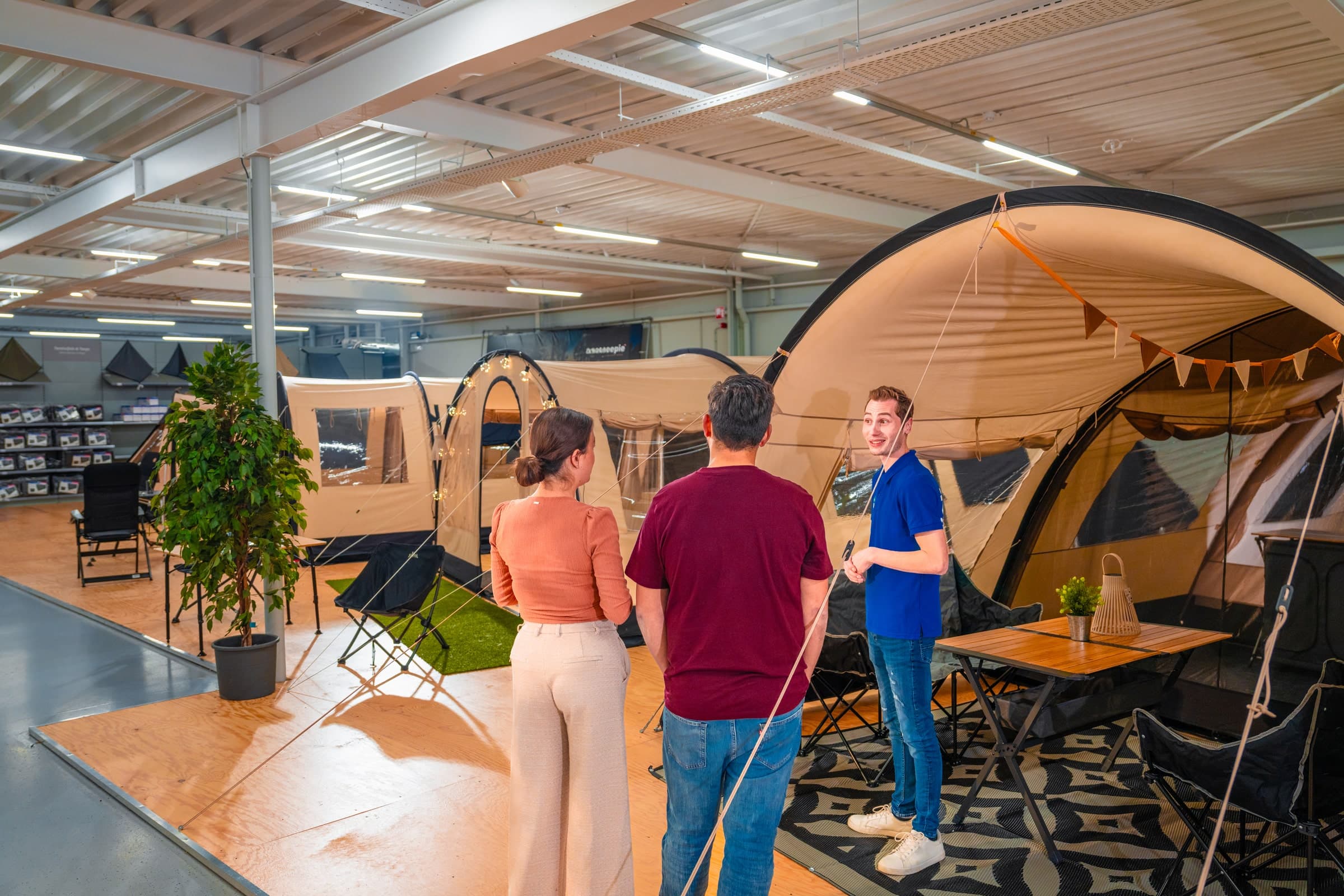 Ruime showroom met opgestelde familietenten, campingmeubilair en decoratieve elementen waar bezoekers de tentopstellingen bekijken