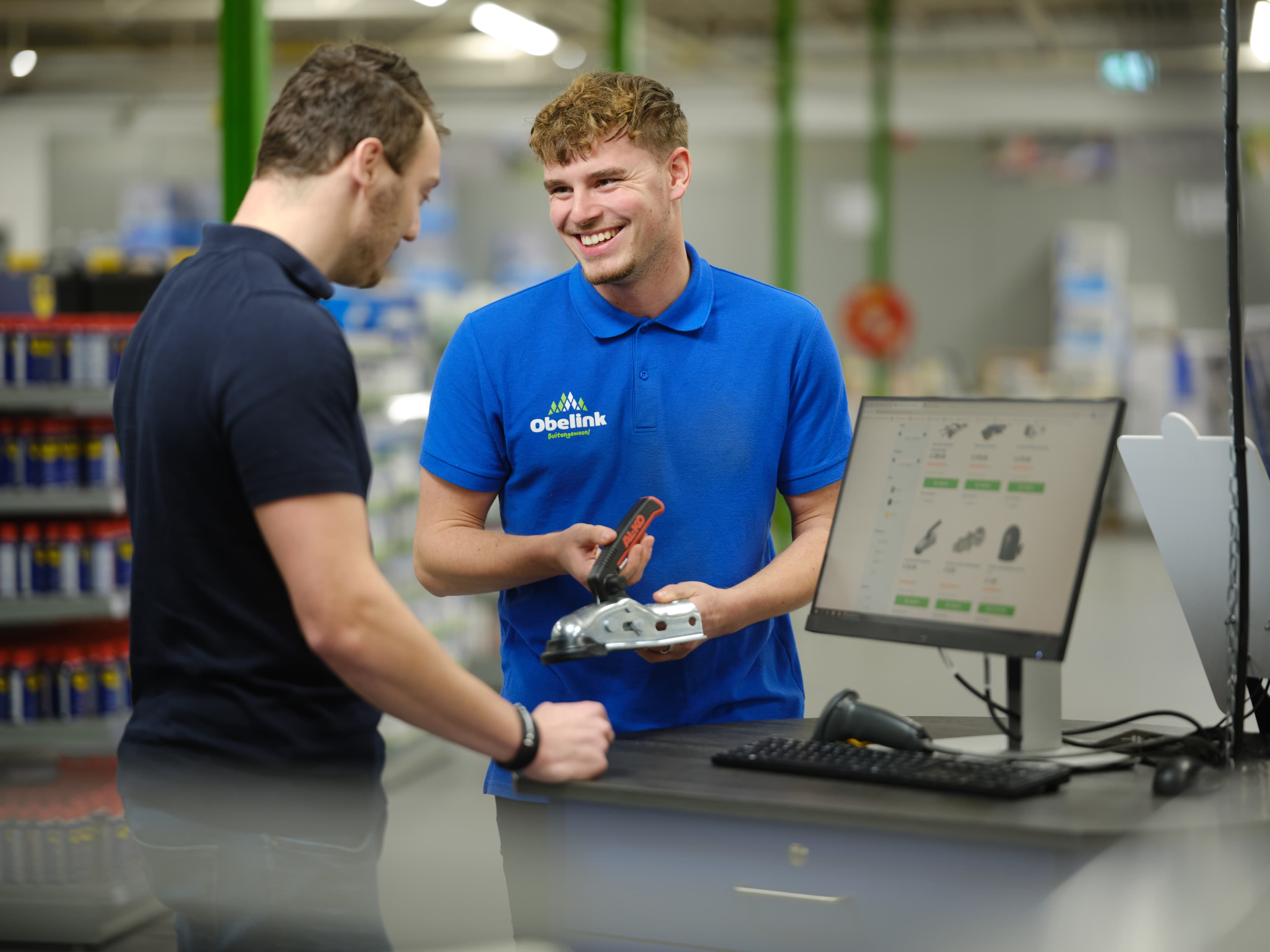 Kom je er niet helemaal uit? Medewerker in blauw Obelink poloshirt toont een metalen bevestigingsonderdeel aan klant bij verkoopbalie met computerscherm waarop producten zichtbaar zijn.