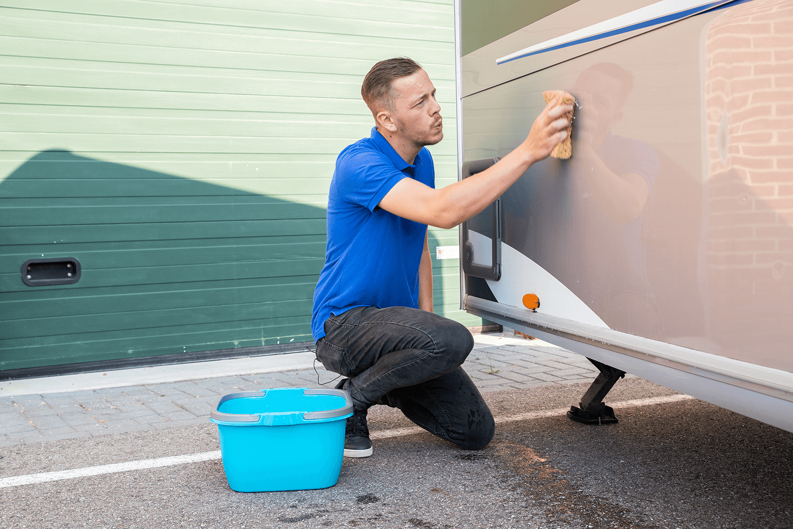 caravan schoonmaken met spons Man in blauw t-shirt reinigt zijkant van caravan met spons, blauwe emmer water ernaast op oprit