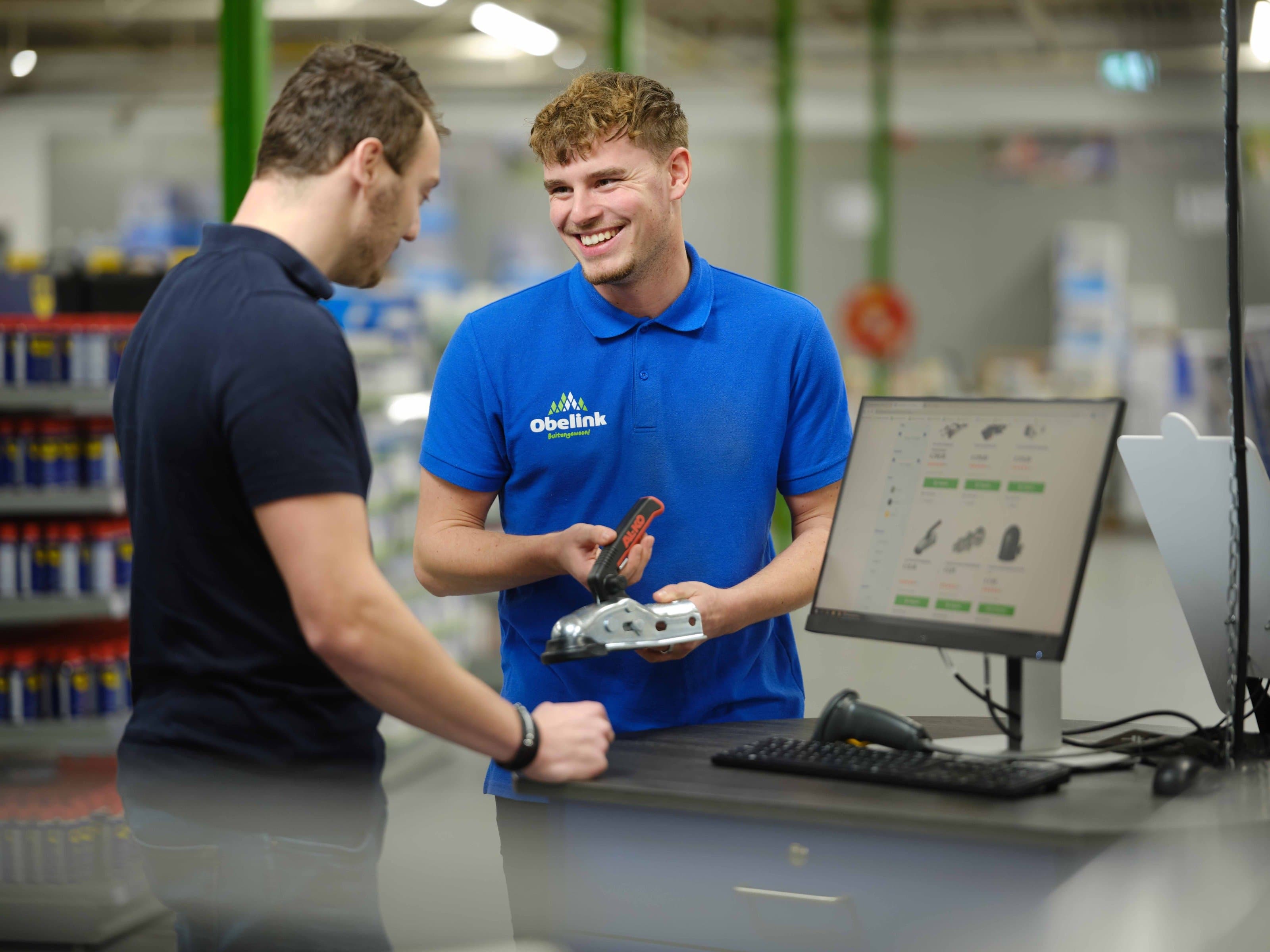 Verkoopmedewerker in Winkel Medewerker in blauw Obelink poloshirt toont een metalen bevestigingsonderdeel aan klant bij verkoopbalie met computerscherm waarop producten zichtbaar zijn