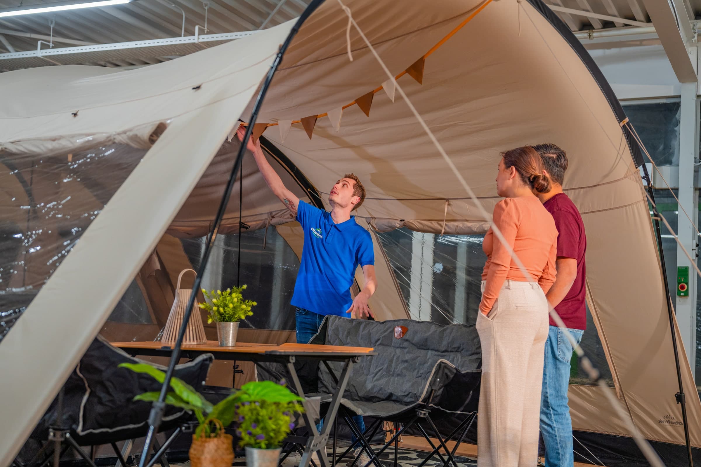 Medewerker in blauw shirt demonstreert een grote familietent aan klanten in een winkel, met kampeertafel en stoelen binnen de opgezette tent
