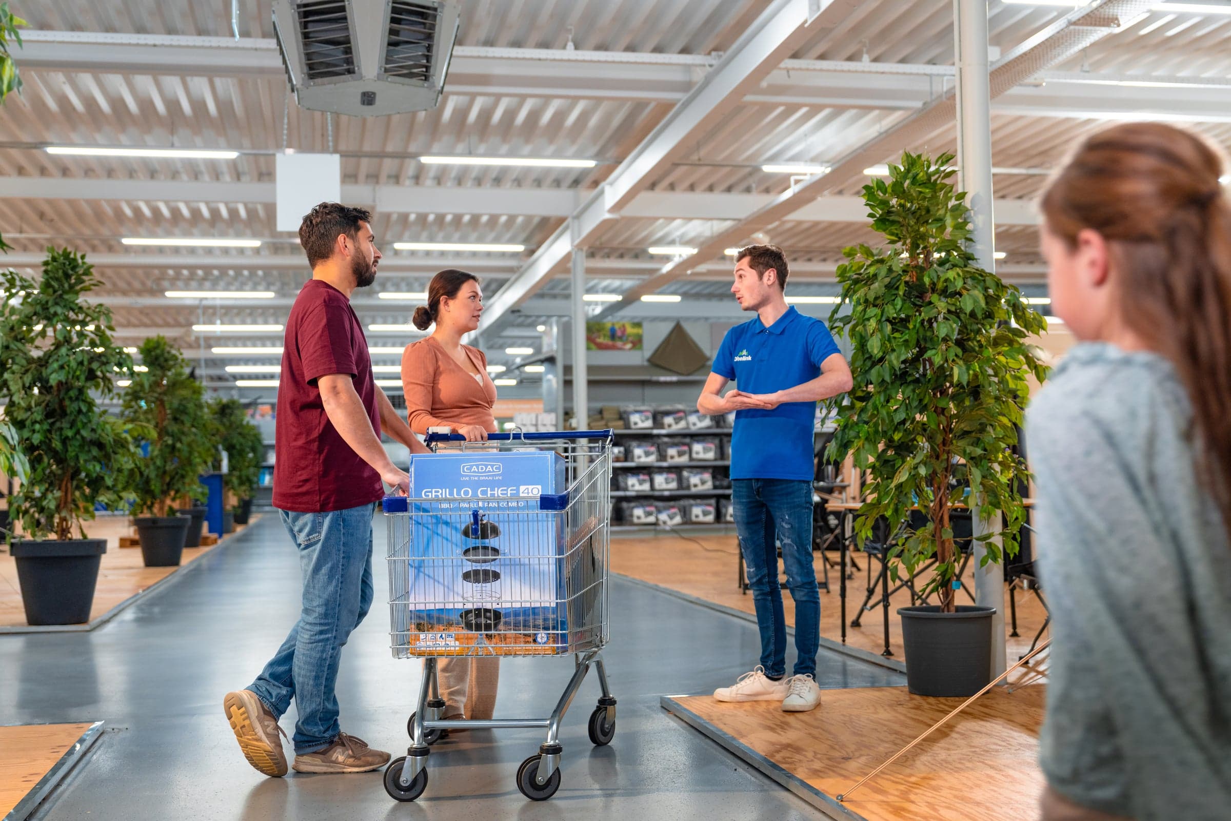 Klanten met winkelwagen en Cadac Grillo Chef 2.0 barbecue krijgen advies van medewerker in blauwe polo in ruime winkel met groene planten
